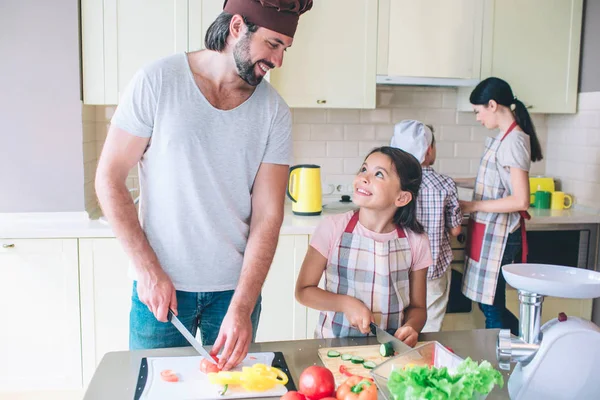 Ευτυχής μπαμπάς στέκεται με την κόρη του και κόβει λαχανικά. Φαίνονται σε κάθε άλλο και χαμόγελο. Μαμα γιος συνεργάζεται πίσω τους στο φούρνο. — Φωτογραφία Αρχείου