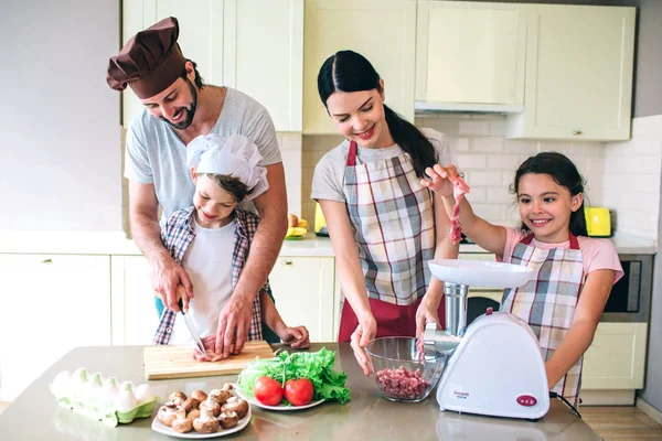 Four members of family are standing together. Guy works with his son. They cut meat. Small girl is playing with piece of it. She wants to put it into meat grinder. Woman holds bowl.