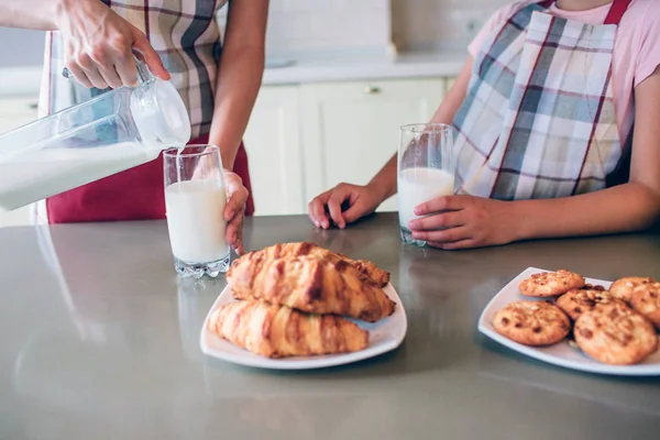 Κομμένα θέα μητέρες χέρια ρίχνει το γάλα σε φλιτζάνι. Αυτή κρατά με το ένα χέρι. Κοπέλα κάνει το ίδιο πράγμα. Υπάρχουν διαφορετικοί τύποι ρολά στο τραπέζι. — Φωτογραφία Αρχείου