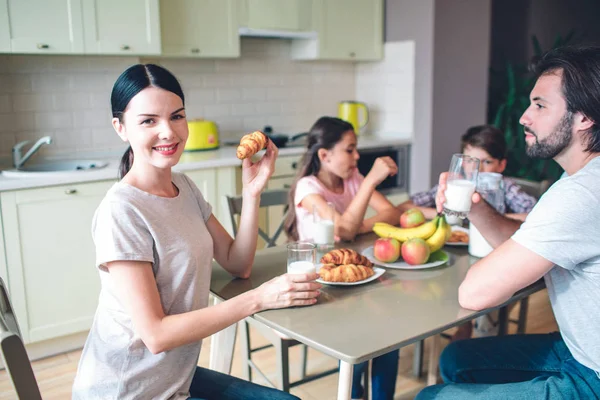 Family are sitting together at table. Woman looks on camera. She holds roll in hand. Other members of family are eating and talking. — Stock Photo, Image