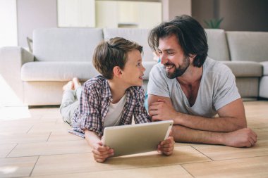 Çocuk ve adam yerde yatıyorlar ve birbirlerine bakıyorlar. Gülümsüyorlar. Çocuğun elinde beyaz dizüstü bilgisayar var. Birlikte vakit geçiriyorlar..