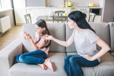 Kız kanepede oturan ve telefon elinde tutan. Annesi çocuk ile tartışıyor. Telefonda işaret eder. Kız üzgün.