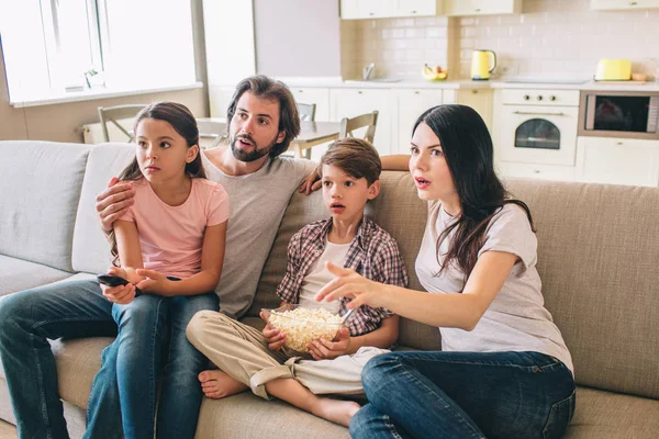 Amazed family sits on sofa and watches TV. They look amazed. Girl sits on daddys lap. Boy has bowl of popcorn. Woman eats this food.