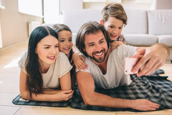 Fammily are lying on the floor. Guy has phone in hands. He look at it and taking selfie of family. Everyone is posing. — Stock Photo, Image