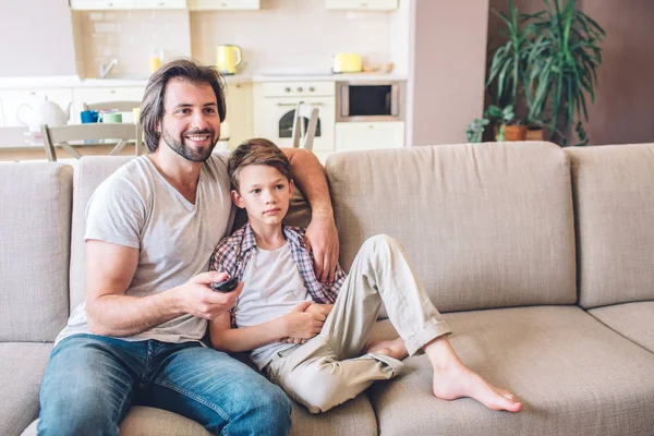 A kép az apa és fia együtt ül a kanapén. Srác tartja a Tv távirányító. Ő használ ez. Fiú hajol az apja. Egyenesen előre néznek. — Stock Fotó