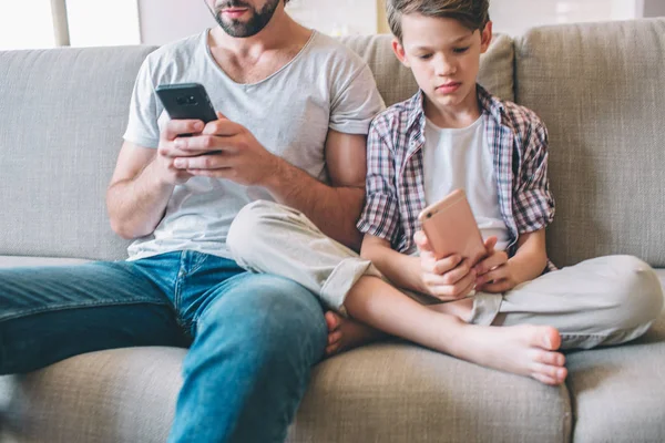 Vágott férfi és fiú ül a kanapén, és nézi a telefon van a kezében. Gyermek ül a lábak corssed. — Stock Fotó