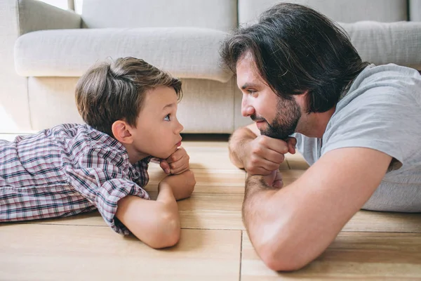 Un hombre y un niño serios y concentrados están tumbados en el suelo y mirándose. Ellos sostienen sus cabezas en manos y puños . —  Fotos de Stock