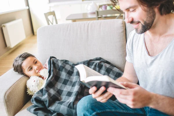 En bild av pappa sitter på kanten av soffan och håller en bok. Han läser den. Flickan är liggande under filt och lyssnar. Killen ler. — Stockfoto