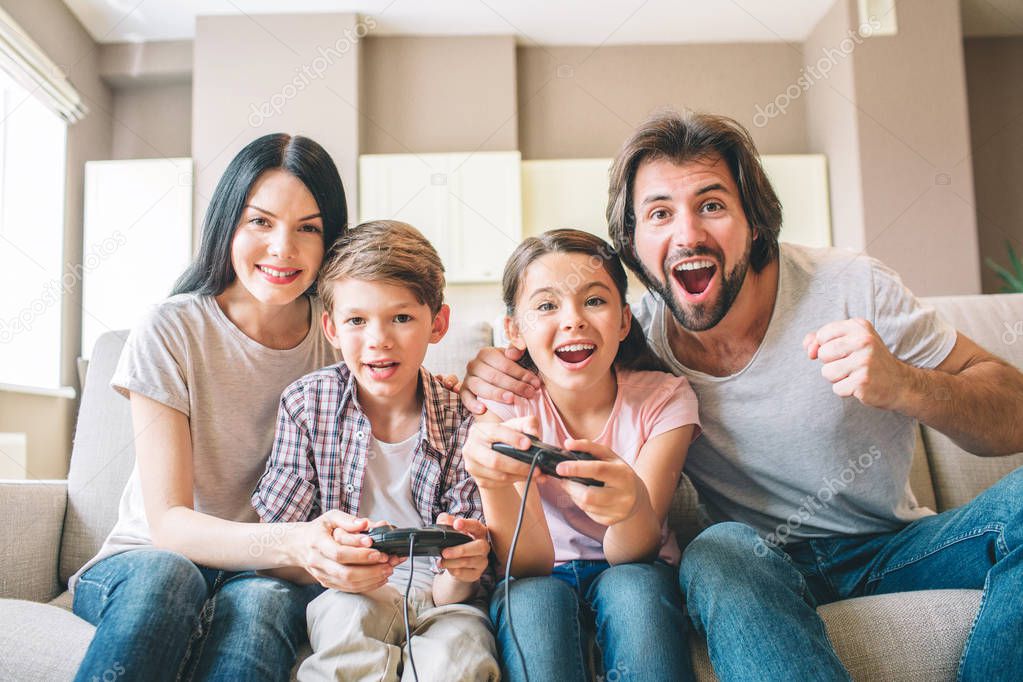 Amazing family sits on sofa. Children are playing on xbox. The game is intense. Guy rejoys. Girl is smiling as well. Mother helps to play her son.