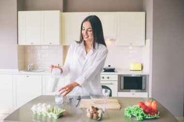 Mutfakta ayakta ve cam fincan süt dökme güzel kızın resmi. O bakıyor ve gülümsüyor. Kız konsantre