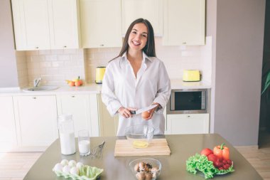 Hoş ve güzel kız tablo ve posese duruyor. O kameraya bakıyor ve gülümsüyor. Kız yumurta ve bıçak elleri. O büyük cam kase onları tutun.