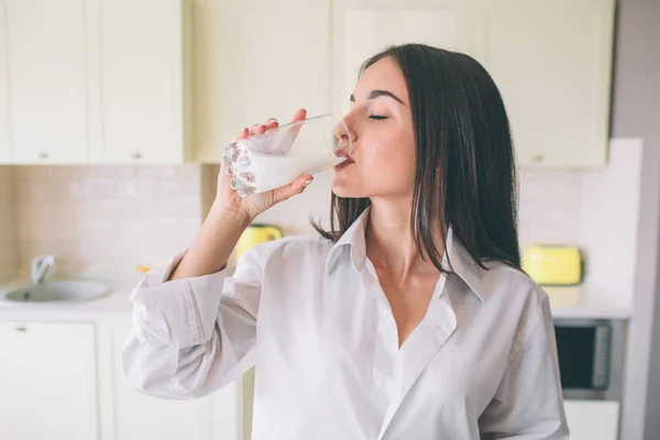 En bild av gorgeus flicka står i köket och dricka mjölk från glas cup. Hon är att hålla ögonen stängda. Kvinnan bär vit skjorta. — Stockfoto