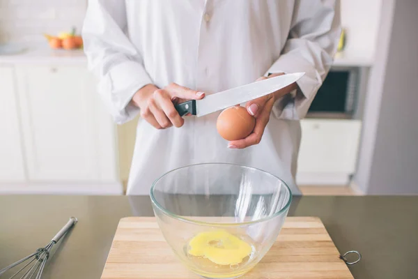 Flicka i vit skjorta står på bordet och håller kniven ovan ägg. Hon kommer att bryta den. Det finns några ägg i stor skål. Det står på träskiva. — Stockfoto