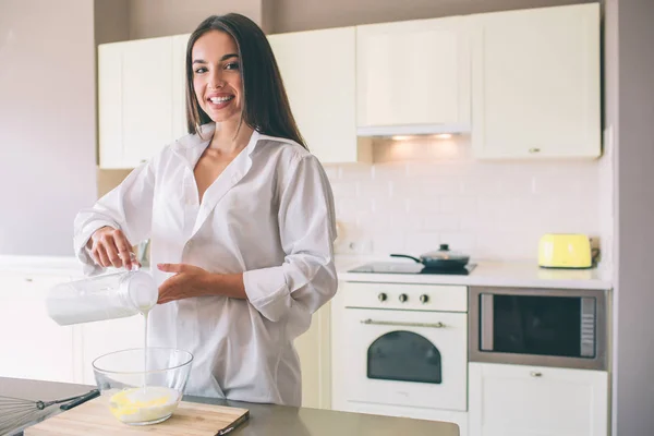 Mujer positiva se para en la cocina y sonríe. Ella está vertiendo leche en un tazón de vidrio grande. Lleva camisa blanca. . —  Fotos de Stock