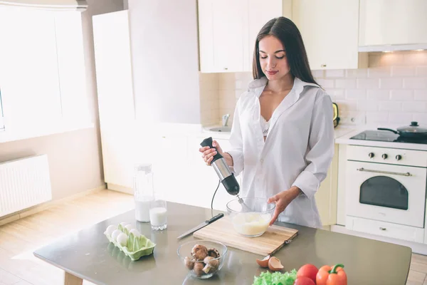 Imagem íntima da menina bonita fica na cozinha e cozinha. Ela está misturando leite e ovos. Há um sutiã branco debaixo da camisa dela. . — Fotografia de Stock