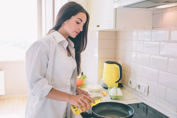 Brunettte fica no fogão e derramando óleo de girassol na frigideira. Ela vai cozinhar. Menina parece séria e concentrada . — Fotografia de Stock