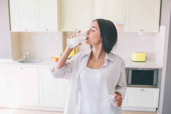 Menina atraente e incrível fica na cozinha à mesa e beber leite. Ela mantém os olhos fechados. Menina aprecia o momento . — Fotografia de Stock