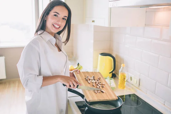 Chica positiva y hermosa se para en la estufa y mirando a la cámara. Ella sonríe. Chica está poniendo setas en sartén . —  Fotos de Stock