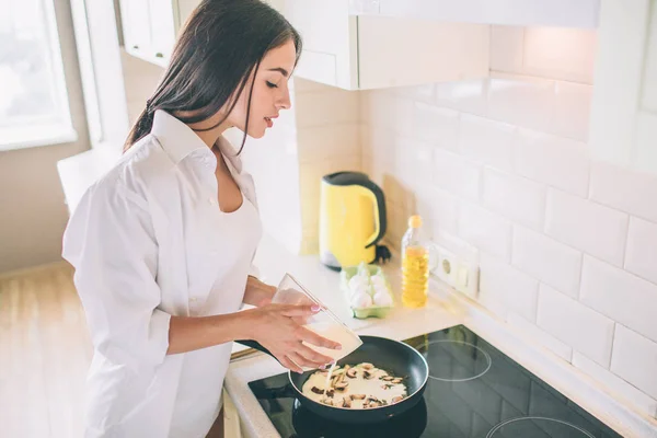 Mulher bonita está cozinhando café da manhã. Ela está derramando ovos misturados e leite em frigideira com cogumelos. A rapariga está a cozinhar o pequeno-almoço. Ela faz isso com cuidado . — Fotografia de Stock