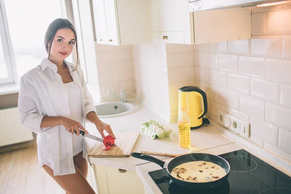 Υπέροχο κορίτσι είναι κοπής ντομάτα σε κομμάτια. Αυτή μαγειρεύει πρωινό. Υπάρχουν αυγά με mushroms τηγάνισμα σε τηγάνι στη σόμπα. Κοιτάζει την κάμερα και χαμόγελα. — Φωτογραφία Αρχείου