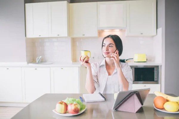 Menina calma e pacífica senta-se à mesa e fala ao telefone. Ela olha para a esquerda. A rapariga tem um copo de sumo na mão. Há tablet, caderno e pratos com frutas na mesa . — Fotografia de Stock