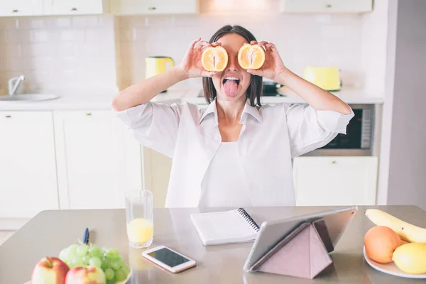 Bonita y divertida foto de una chica escondiendo sus ojos con trozos de naranja. Ella está emocionada. Hay una tableta. teléfono, portátil, vaso de jugo y platos con fruta en la mesa . —  Fotos de Stock