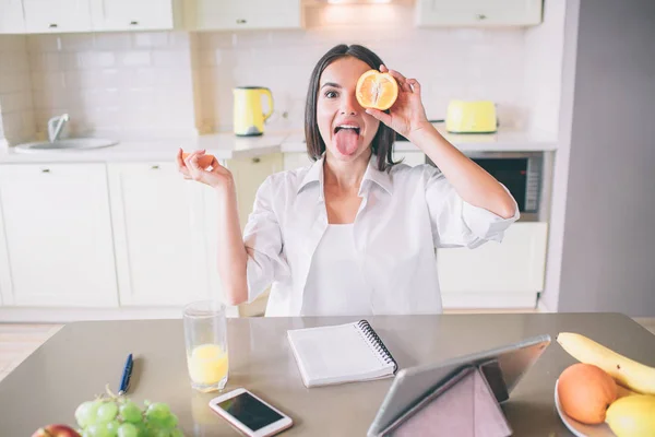 Zabawna dziewczyna bawi się z owoców. Ona hold kawałek pomarańczy przed oczami i pokazuje jej język. W ręku trzyma anotheer część owocu. Dziewczyna siedzi w kuchni. — Zdjęcie stockowe