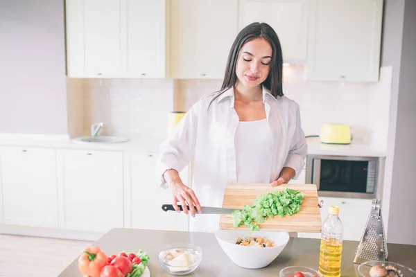Προσεκτική και όμορφος κορίτσι στέκεται στην κουζίνα και το μαγείρεμα σαλάτα. Βάζει cutted μαρούλι σε μπολ με ανάμεικτα υλικά. — Φωτογραφία Αρχείου
