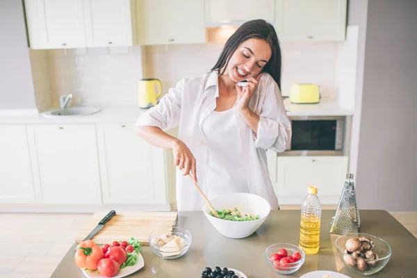 La chica bonita está hablando por teléfono mientras está cocinando. La mujer está mezclando ingredientes para ensalada. Ella usa cuchara de madera para eso. . —  Fotos de Stock