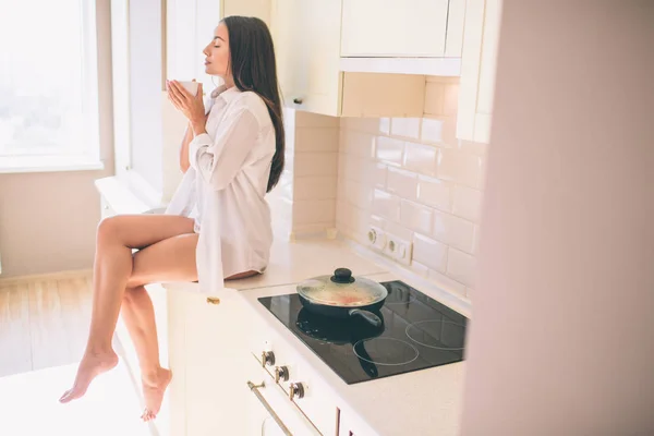 Bella ragazza con corpo stupendo è seduto sul tavolo vicino stufa e in attesa di cibo per essere cucinato. Sta tenendo le gambe incrociate . — Foto Stock