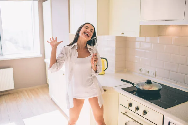 Happy girl is dancing and singing. She has a lot of fun while food is cooking in pan. She holds mixer in hand. Girl pretends it is microphone. — Stock Photo, Image