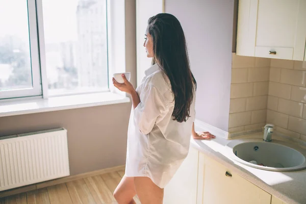 Hermosa e impresionante chica se para en la cocina y mirar a la ventana. Ella es fuerte y bien construida. Chica está posando . —  Fotos de Stock