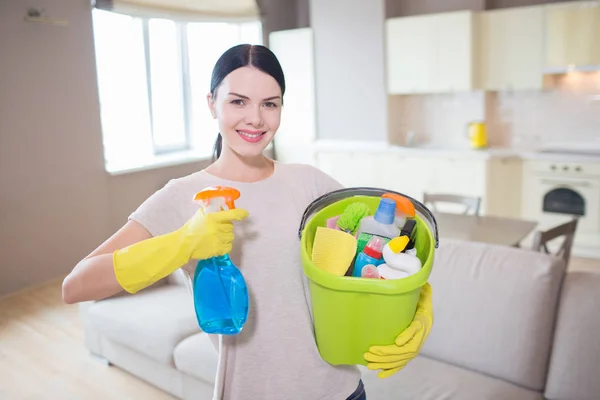 Krásná a pozitivní žena stojí a drží modrý sprej a kýbl s čištěním zařízení v rukou. Vypadá a představuje na kameru. — Stock fotografie