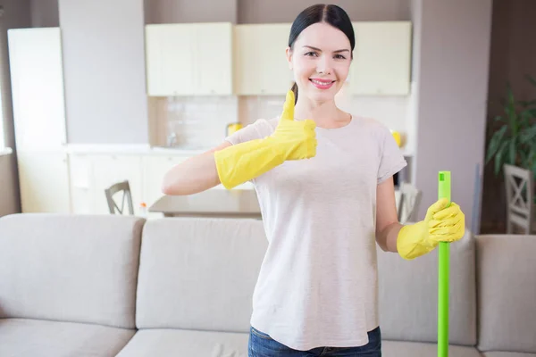 Dziewczynka stoi w mieszkaniu i pokazuje jej wielki kciuk. Kobieta nosi rękawice żółty i przytrzymaj zielony trzymać lewą ręką. Ona jest pozytywne. — Zdjęcie stockowe