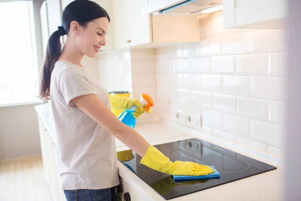 Nice and positive brunette stands in front of stove and cleans it with rag. Also she holds blue liquid spray in left hand. Girl wears yellow gloves. — Stock Photo, Image