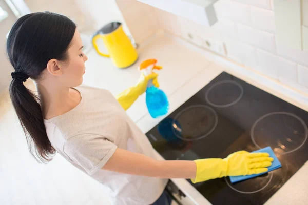 Dark-haired girl stands in front of dark stove and cleans it from dirt. She uses spray and blue rag for that. Young woman is doing that careful.