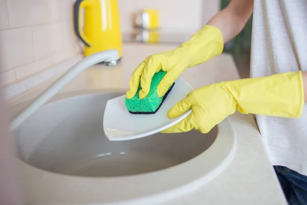 A picture of humans hands washing the dishes. People uses green sponge and wears yellow gloves.