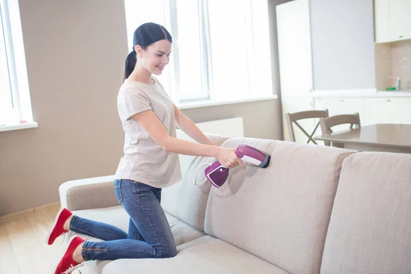 Pěkné a pozitivní dívka používá malý vysavač na pohovce. Ona je čištění. Dívka stojí na kolena na pohovce. — Stock fotografie