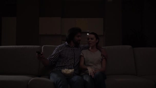 Lovely couple sits in dark and watch TV. Guy has bowl of popcorn on knees. Girl wants to get remote control. Guy doesnt want to give it but he smiles. Girl pretends she is upset but continue watch. — Stock Video