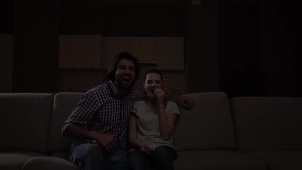 Couple sits on sofa and wathes movie. Guy has bowl with popcorn on knees. They start to laugh. People are watching comedy. Girl is pointing forward and looking at guy. — Stock Video