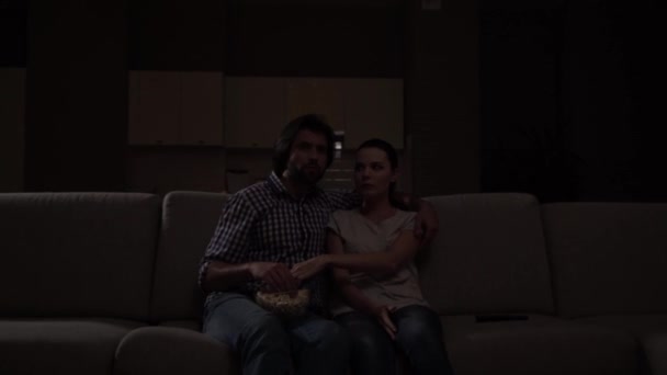 Man and woman sit on sofa and watching movie. They eat popcorn. Guy holds bowl with junk food on knees. Girl got scared. She coveres mouth with hand. Guy protects her. — Stock Video
