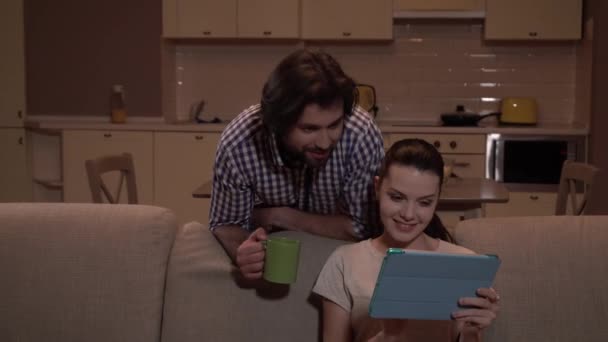 Girl sits on sofa and uses tablet. She looks at it and shows to guy. He stands behind sofa and look at tablet. Man holds cup. Girl is amazed and excited. — Stock Video