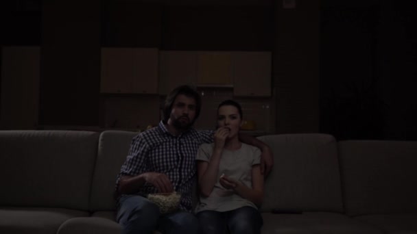 Homem e mulher sentam-se juntos e assistem TV. Um tipo a abraçar uma rapariga. Eles comem pipocas. O filme está muito animado. Eles estão concentrados . — Vídeo de Stock