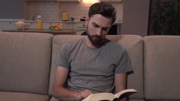 Бородатый парень сидит на диване и читает толстую книгу. Он переворачивает страницы. Затем человек смотрит влево и продолжает читать. . — стоковое видео