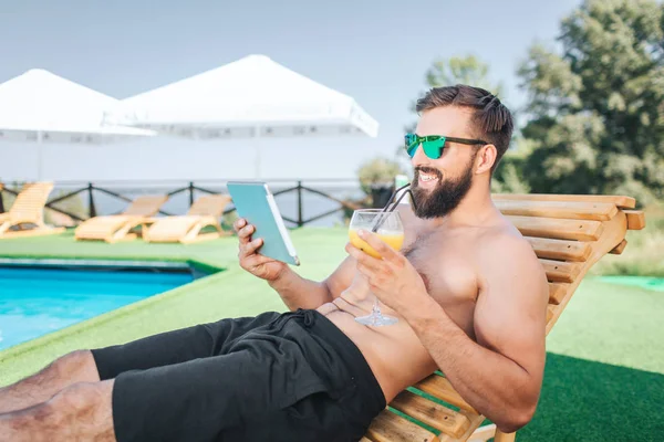 El tipo relajado y feliz se sienta en la cama de sol y sonríe. Él sostiene la tableta y la mira. El hombre usa gafas de sol. Tiene un cóctel en la mano izquierda. Está sentado junto a la piscina. Guy está satisfecho. . — Foto de Stock