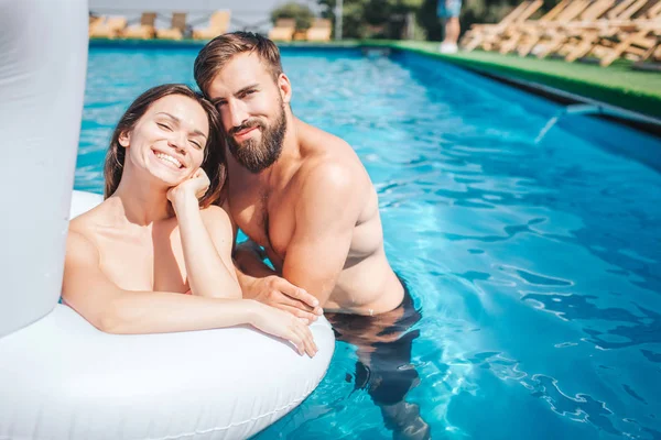 Nce y los jóvenes positivos están nadando en la piscina. La chica lo hace en colchón de aire. El tipo está nadando en el agua y se inclina hacia la chica. Parecen felices. . —  Fotos de Stock