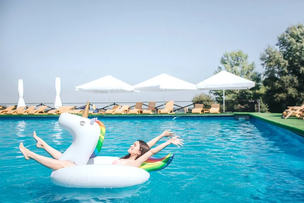 Happy and satisfied girl is lying on air mattress in the middle of swimming pool. She keeps hands and legs in air. Girl is relaxing. She has some rest. — Stock Photo, Image