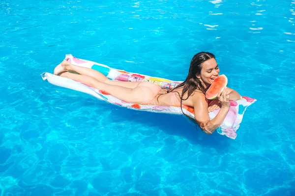 Happy girl is lying on air mattress and eat piece of watermelon. She smiles. Girl is in swimming pool. — Stock Photo, Image