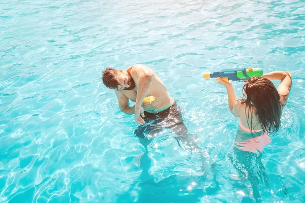 Imagen divertida de pareja jugando en la piscina. La chica está disparando a un tipo de pistola de agua. Intenta defenderse. Se divierten mucho. . —  Fotos de Stock