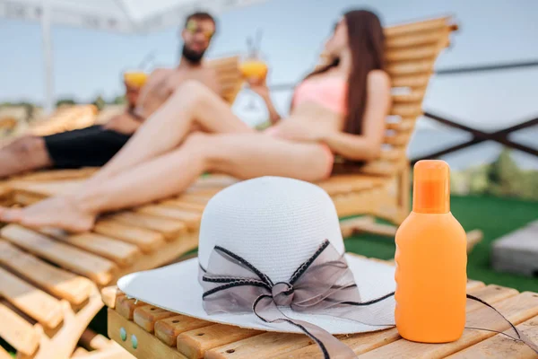 A picture of orange bottle and hat lying on small wood table. Couple is sitting behind it. They look at each other and hold glasses og cocktails in hands
